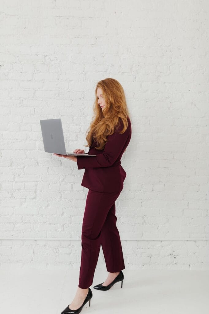 Cheryl Ann Barnes standing against a white brick wall in a burgundy suit, typing on an open laptop she holds in her arms