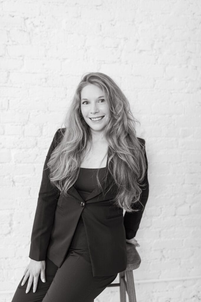 Cheryl Ann Barnes in a black blazer, black-and-white headshot, leaning against a stool and smiling at camera
