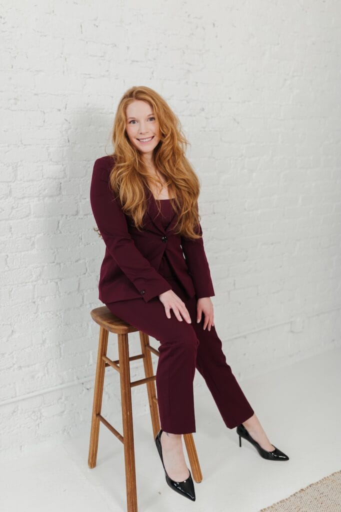 Cheryl Ann Barnes seated on a wooden stool in a burgundy suit, smiling at camera against a white brick wall