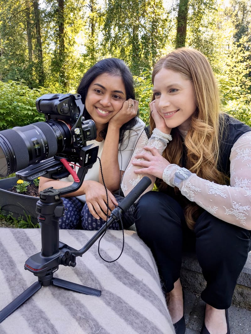 Cheryl Ann and a colleague resting beside a professional video camera during an outdoor shoot in a green garden