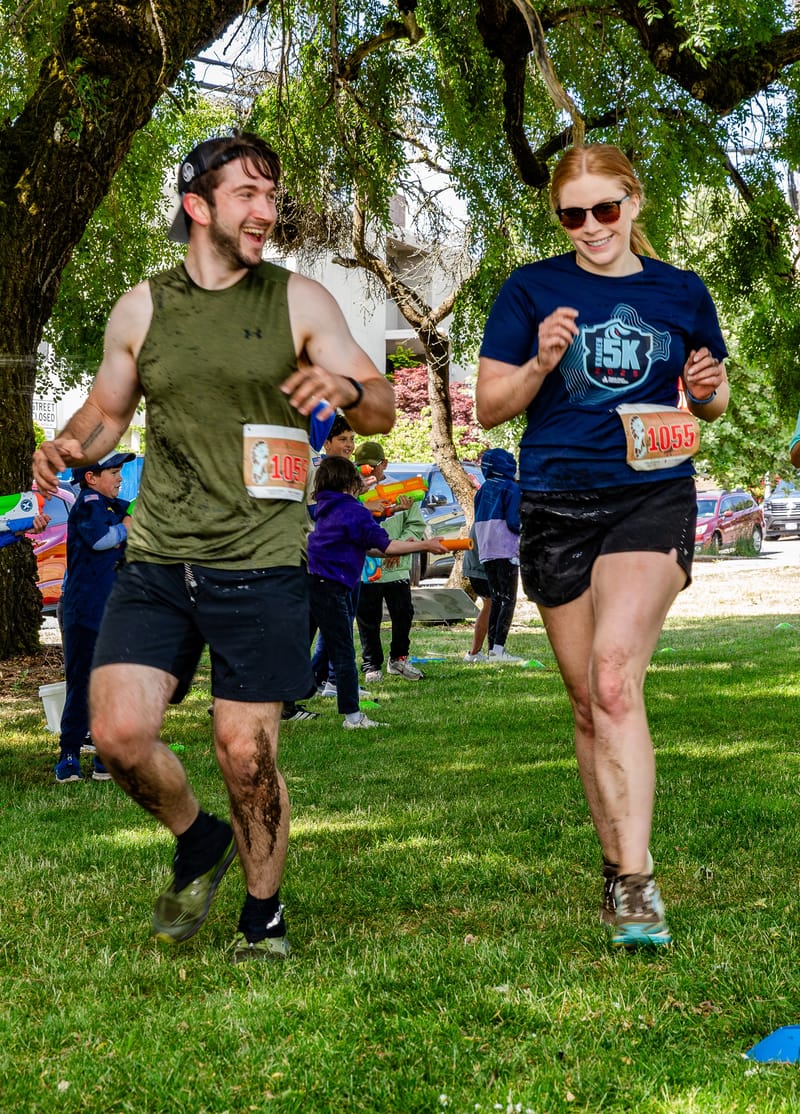 Cheryl Ann and a fellow runner finishing a community 5K through a sunny park, with kids spraying water guns along the course