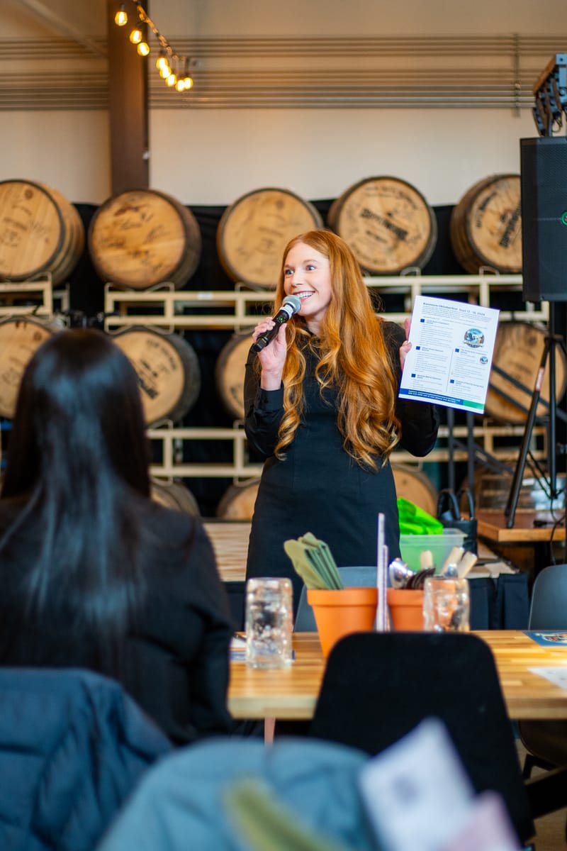 Cheryl Ann speaking into a microphone and holding a Kenmore Oktoberfest flyer in front of barrels at a brewery